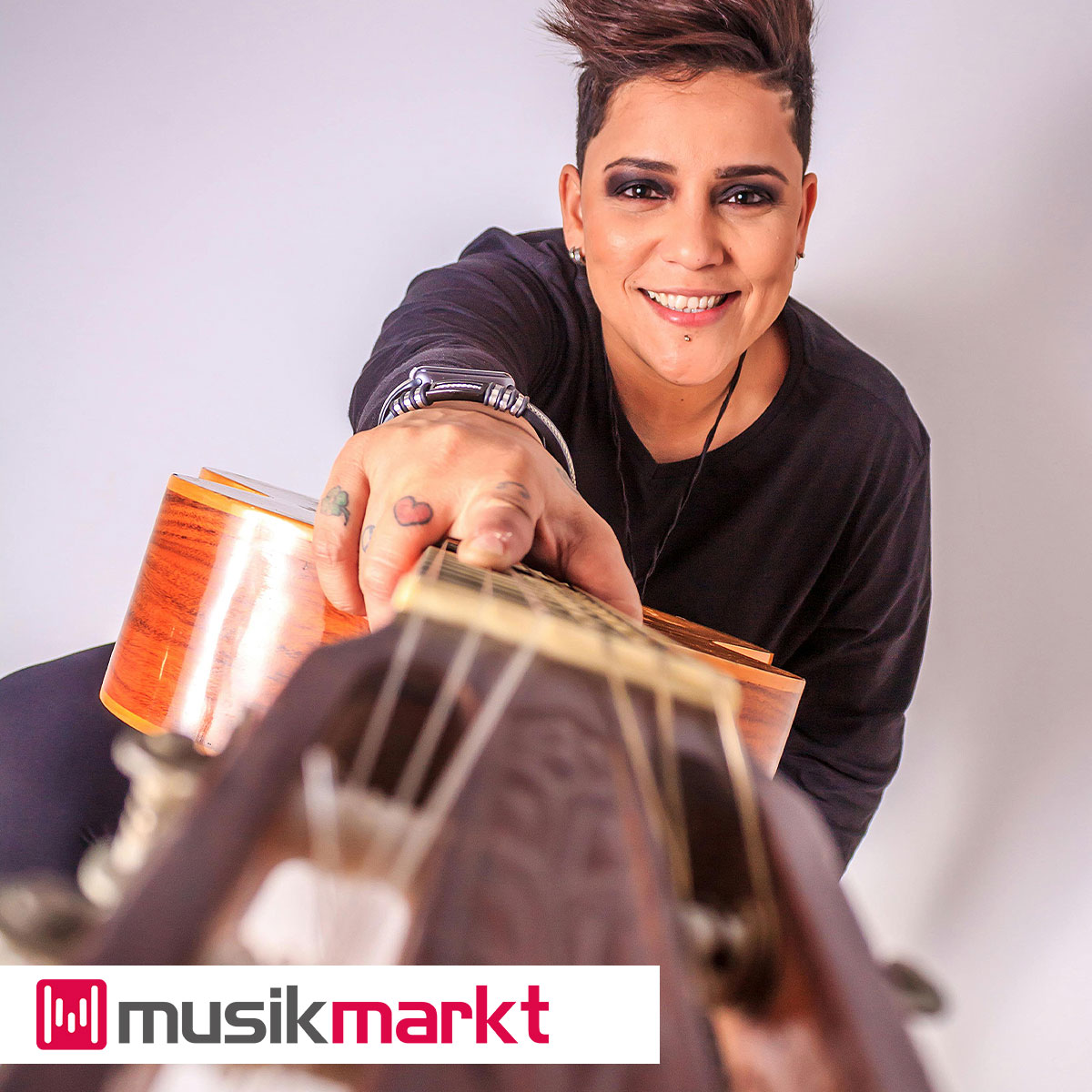 Frau mit kurzen rotbraunen Haaren und dunklem Pullover, die lächelnd eine Gitarre auf den Betrachter zuhält.