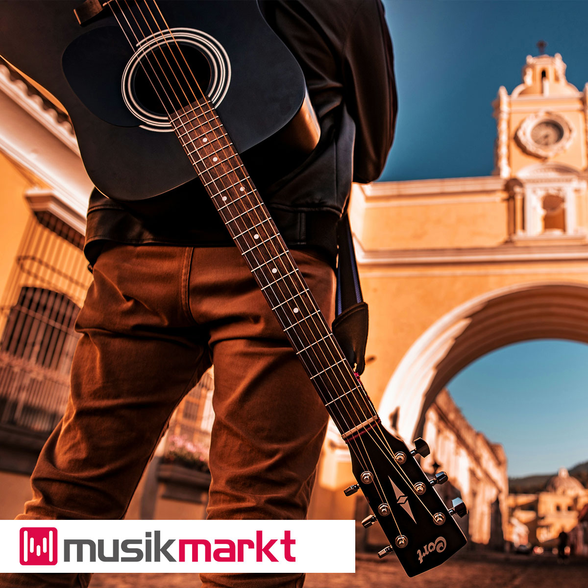 Person mit einer schwarzen Cort Gitarre auf dem Rücken steht bei blauem Himmel vor einem Torbogen.